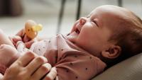 Mother plays with young smiling baby who is resting on her legs.